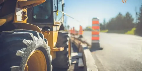 Close-up view of road construction machinery applying advanced Polymer Modified Bitumen (PMB) on a highway under development, with safety barriers and blurred cityscape in the background